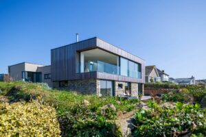 Modern two-story house with large glass windows and double glazed doors, a mix of wood and stone exterior, surrounded by greenery under a clear blue sky.