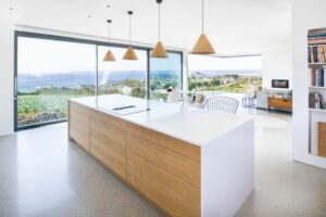 A modern kitchen features a large island, wooden cabinets, and pendant lights, with double glazed sliding glass doors offering a breathtaking view of the ocean. In the background, a dining area invites you to enjoy meals with serene vistas.