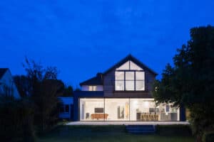 A modern two-story house with large windows is illuminated at night, showcasing an open-plan living area with sleek balustrades, a pool table, and a dining table.