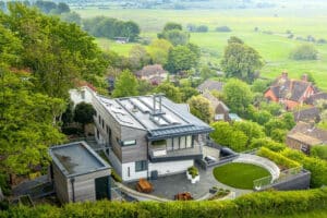 A modern house with a flat roof and sleek balustrades, surrounded by trees and greenery, overlooks a residential area and expansive fields.