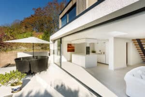 Modern home with open sliding aluminium doors leading to a patio. Inside is a white kitchen; outside, a round dining table with an umbrella. Bright sunlight illuminates both areas.