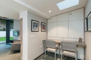 A small minimalist workspace with two chairs and a desk. The white walls feature three framed pictures, while a teapot and mugs rest to the right. An aluminium bifold glass door elegantly opens to the outside world.