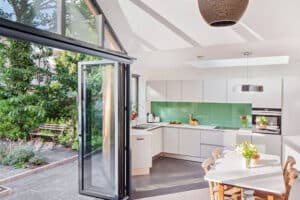 Modern kitchen with white cabinets, a green backsplash, and a dining table. A glass door opens to a garden with greenery and a wooden bench. A pendant light hangs above the table, casting warmth through the space's elegant glazed gable end.