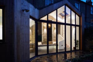 A modern house exterior features large glass windows and wooden cladding, showcasing a glazed gable end. Inside, a lit brick wall and staircase come alive at night, blending warmth with contemporary design.
