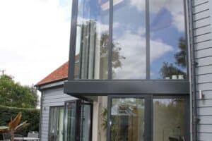A modern house boasting a tall glass extension with a sleek glass roof, complemented by a patio. The exterior includes a table, chairs, and potted plants for an inviting outdoor space.