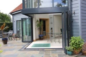 Open glass doors lead from the modern house to a stylish extension with a glass roof, showcasing a patio adorned with outdoor seating and lush plants.