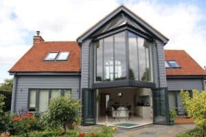 Modern two-story house with large glass windows, gray siding, and a red-tiled roof. The open patio doors lead to a kitchen and garden area, featuring an elegant extension with a glass roof that bathes the space in natural light.