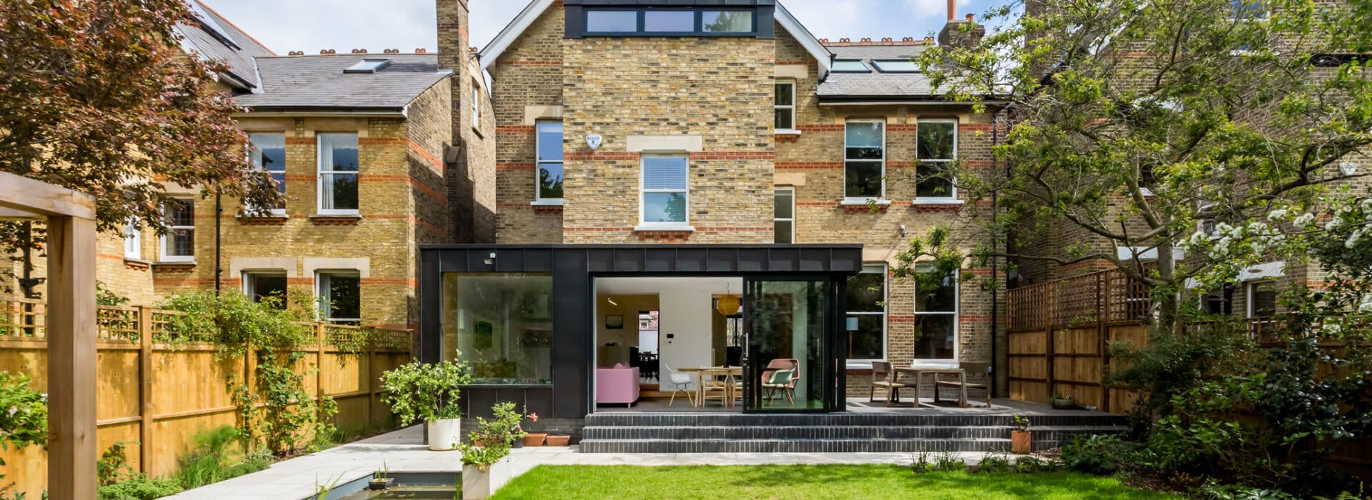 Modern brick home extension featuring expansive glazing systems with black framing, bifold doors opening onto a patio, and integrated roof lights creating seamless indoor-outdoor living.