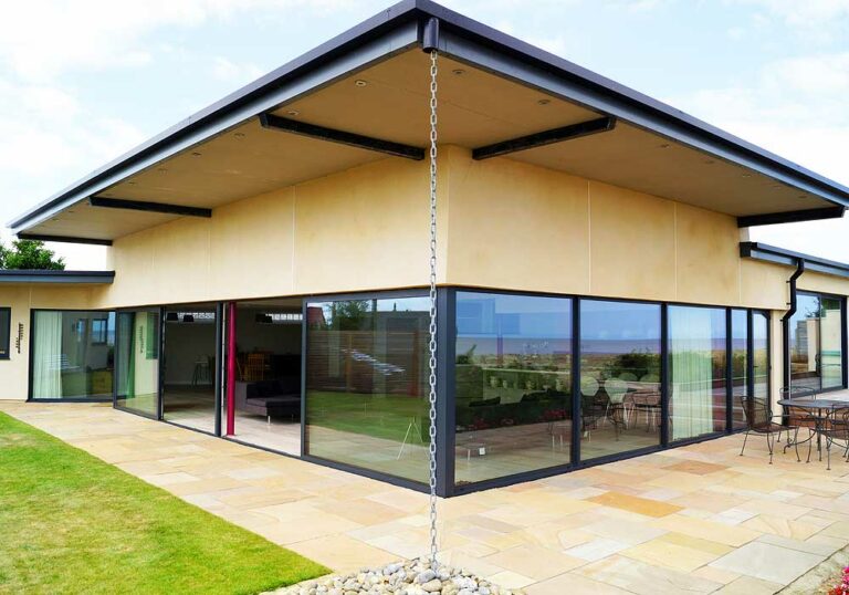 Modern house with large picture windows, a flat roof, and a rain chain at the corner. The patio has stone tiles and outdoor seating.