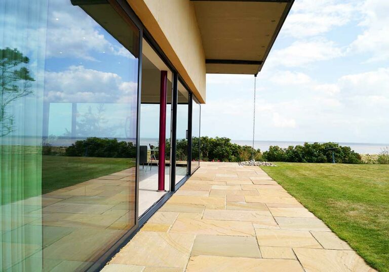 Modern house with expansive picture windows overlooking a grassy yard and distant sea view on a sunny day.