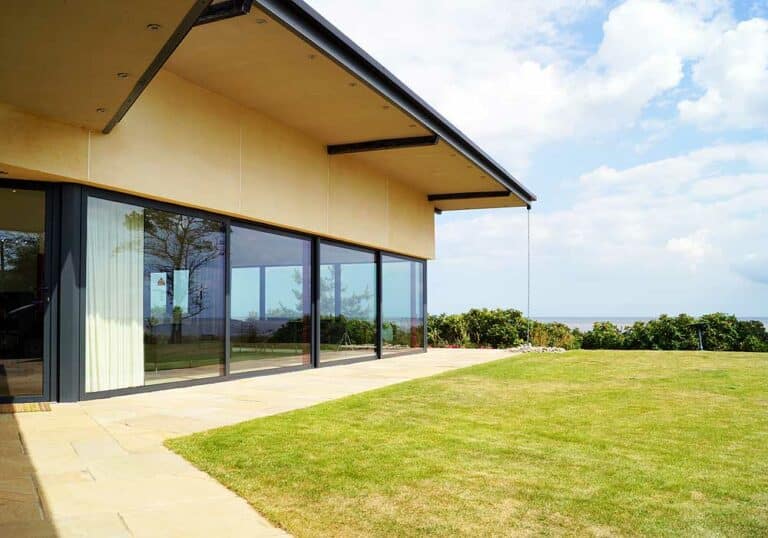 A modern house with expansive picture windows overlooks a grassy lawn and a distant ocean view under a partly cloudy sky.