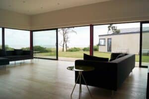 Modern living room with large picture windows overlooking a garden and sea view, featuring black sofas and small round tables.
