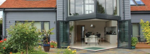 Modern house with a red-tiled roof features large glass doors opening to a patio, offering a glimpse of the kitchen inside. An extension with a glass roof allows natural light to flood the space, while the plant-filled garden enhances its charm.