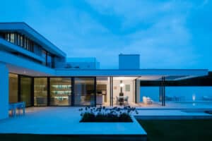 Modern house with large glass windows, flat roof, and outdoor patio area illuminated in the evening. Featuring corner bi-fold doors, the minimalist design flows seamlessly into a garden and pool.