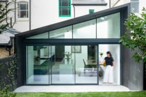 Modern extension with large glass sliding doors and a sloped roof, featuring a visible dining area inside. A person is walking by the door.