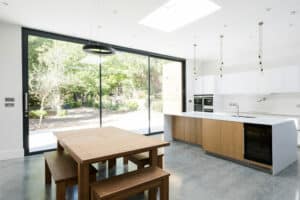 A bright modern kitchen featuring a large glass sliding door, a wooden dining table with benches, an island with a white countertop, pendant lights, and stainless steel appliances.