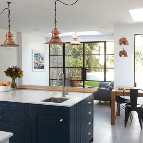 Modern kitchen with navy island, copper pendant lights, and sink. Open layout to dining area features window view through traditional steel frame doors. Decor includes a vase of flowers and wall art that nods to the home's art deco heritage.