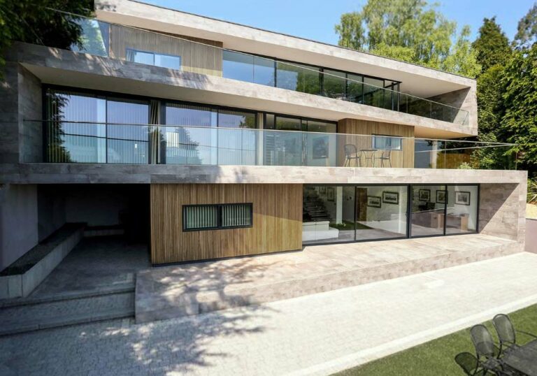 Modern three-story house with large glass windows, balconies, and a wooden facade featuring sleek aluminium sliding doors. Surrounded by trees and a paved pathway, two chairs are visible on the lawn.