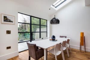 A modern dining room features a white table, eight chairs, and a woven pendant light beneath a skylight. Aluminium heritage doors complement the wooden flooring, enhancing natural light from the large window. A decorative bowl adorns the table.