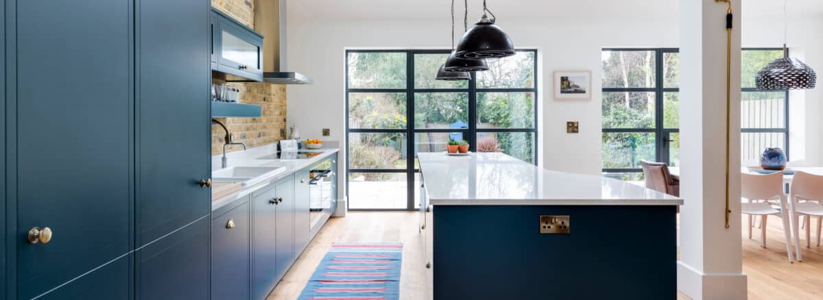 Modern kitchen with blue cabinets, white countertops, and pendant lights over an island. Large windows and sleek aluminium heritage doors provide natural light. A dining area with a pink chair is visible in the background.