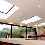 Modern kitchen with expansive roof lights, ceiling fans, and floor-to-ceiling windows overlooking a garden. Artwork adorns the wall, and wine bottles rest elegantly on the counter.