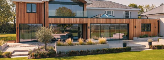 Modern two-story house with a wood and glass exterior, featuring slimline sliding doors and large windows that open to a landscaped garden in the foreground.