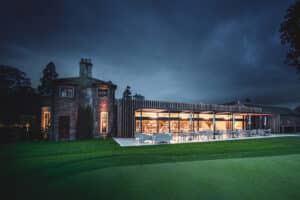 A modern building with large glass windows and a brick section, lit warmly from inside, set against a dark, cloudy evening sky, with a green lawn in the foreground.