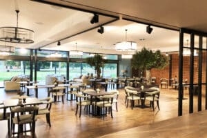 Spacious modern restaurant interior with wooden tables, cushioned chairs, large windows, greenery, and hanging lights.