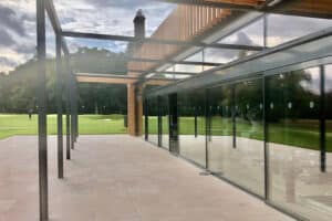 Modern building with large glass windows, reflective surfaces, and an open patio. Wooden beams on the roof, grassy landscape with trees in the background. Cloudy sky.