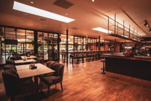 A spacious, modern restaurant with wooden floors, tables, and chairs. The interior features a long bar, large windows, and warm lighting.