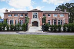 Red brick mansion with multiple windows, centered entrance, and manicured hedges along a paved driveway.