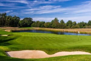 A golf course with a sand trap in the foreground, a pond behind the green, and trees lining the background under a blue sky.