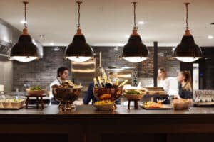 A modern kitchen setting with three people and a central counter displaying dishes of food and bottles in a bowl. Four large hanging lights illuminate the scene.