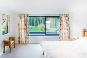 A bright bedroom with a large bifold window, floral curtains, a bed with white bedding, and wooden furniture. Outside, there's a beautiful view of greenery and trees.
