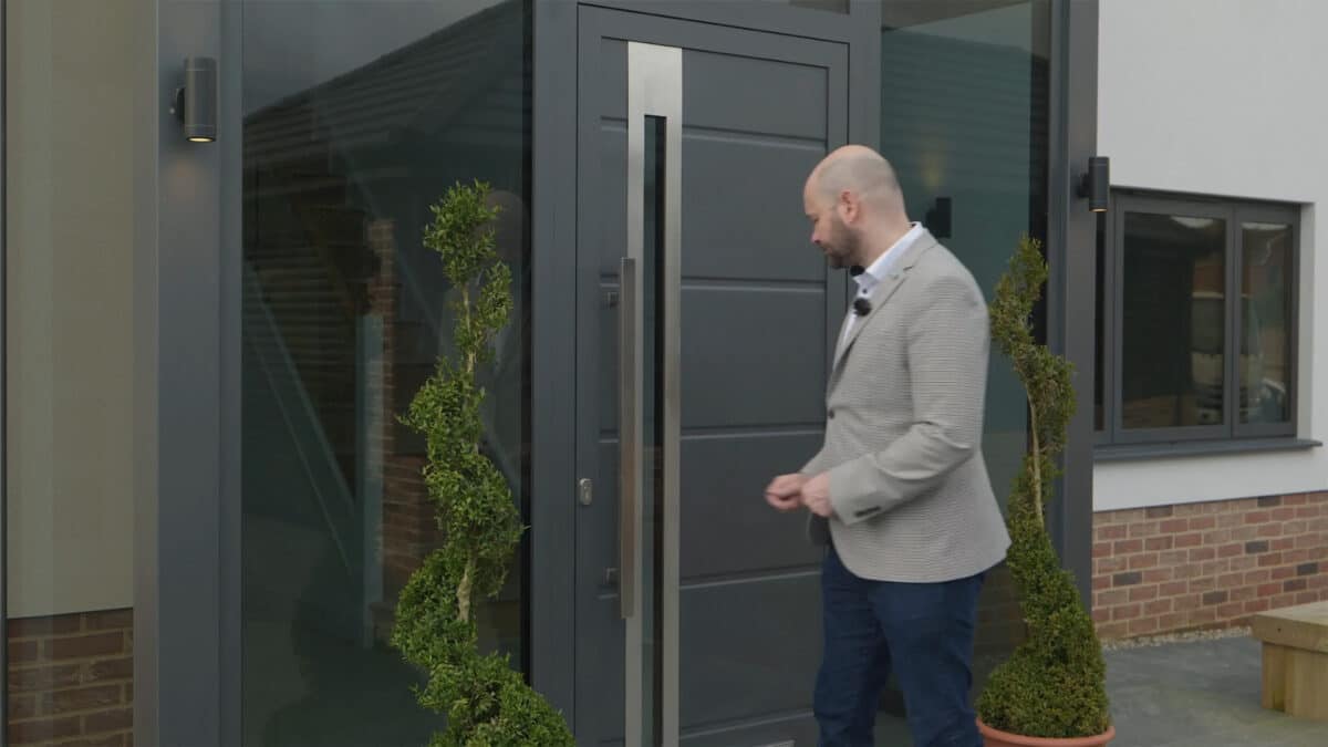 A man in a gray jacket stands in front of a sleek, modern front door, flanked by two spiral-shaped topiaries on either side.