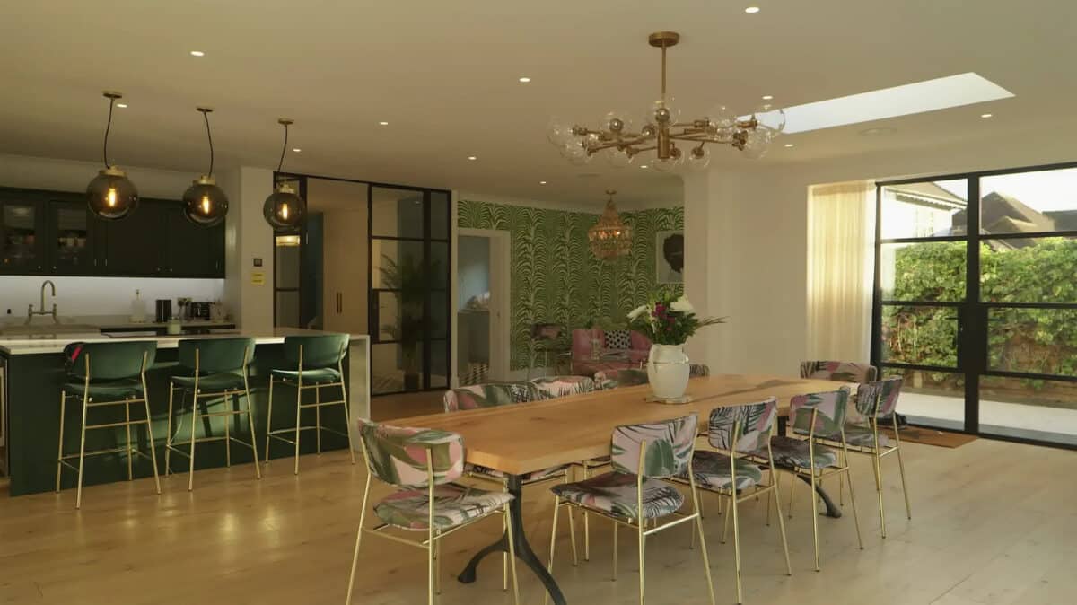 Spacious kitchen and dining area with a long wooden table, decorative chairs, a green-themed accent wall, and modern lighting fixtures. Heritage doors crafted from glass lead to an inviting outdoor garden.