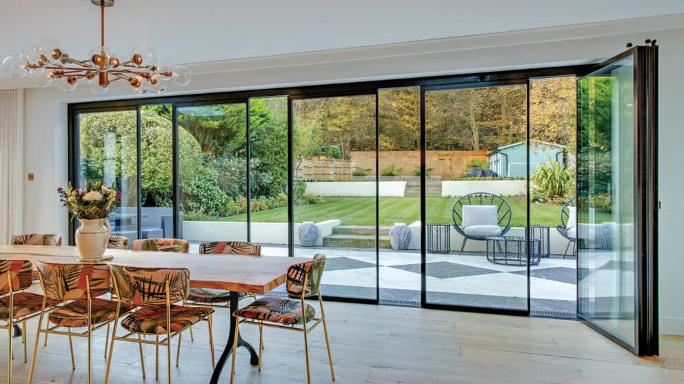 A dining room with a wooden table, colorful chairs, and a modern chandelier. Bifold doors open to a patio with chairs and a garden.