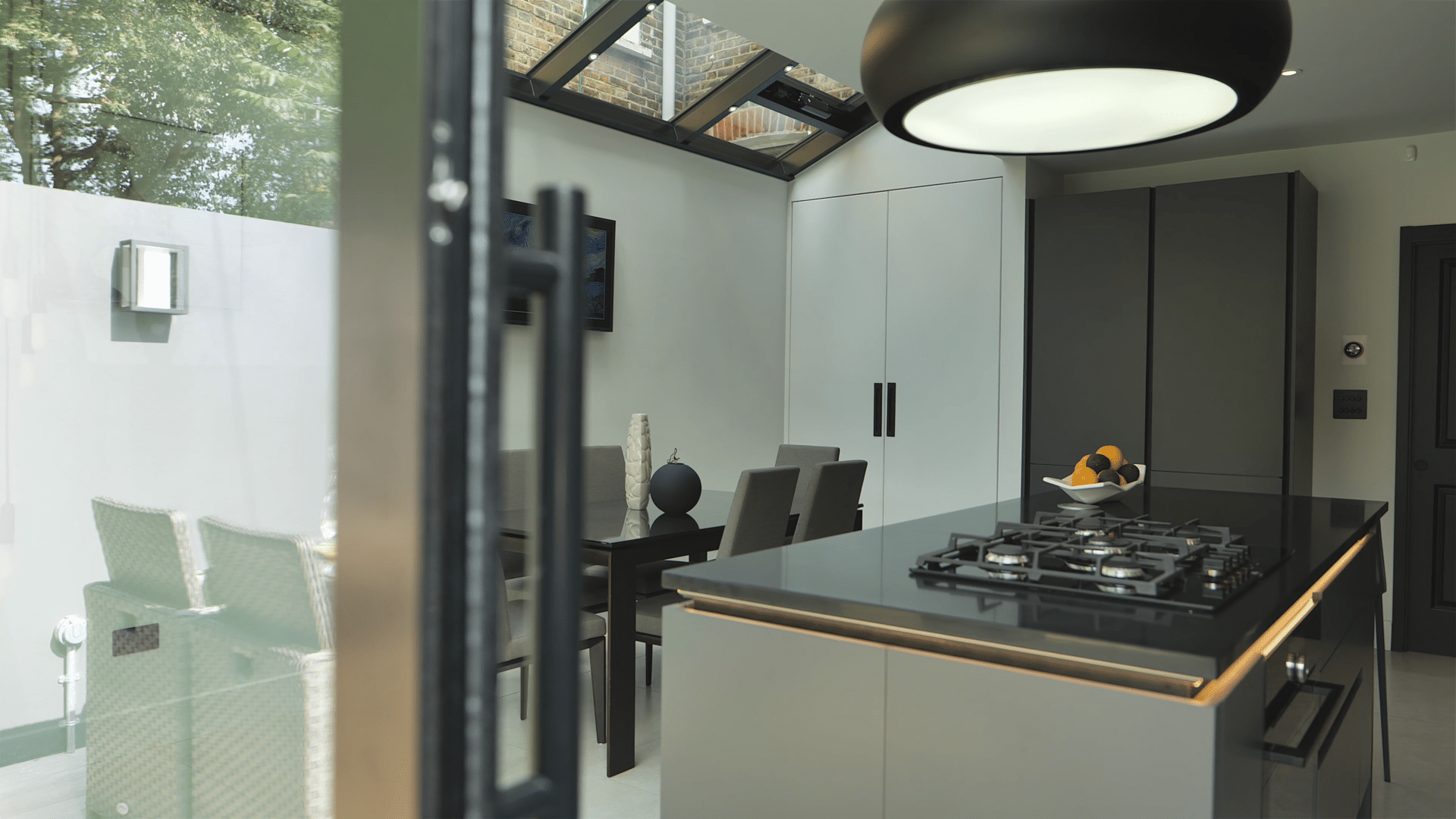Modern kitchen with a glass roof flooded with natural light, featuring sleek black cabinetry, a gas hob, and gold-trimmed black island countertop.