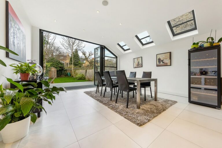 Modern dining room with a large table, six chairs, and skylights. Double glazed bifold doors lead to a garden, allowing seamless indoor-outdoor flow. Floor-to-ceiling windows illuminate the space, which is adorned with indoor plants and artworks.