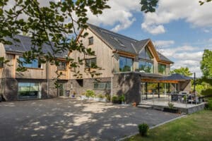 Modern two-story house with wooden accents, large glass windows, and solar panels. Featuring a 4 panel sliding glass door leading to an outdoor patio under a blue sky with clouds. Surrounded by trees and greenery, this home seamlessly blends nature with contemporary design.