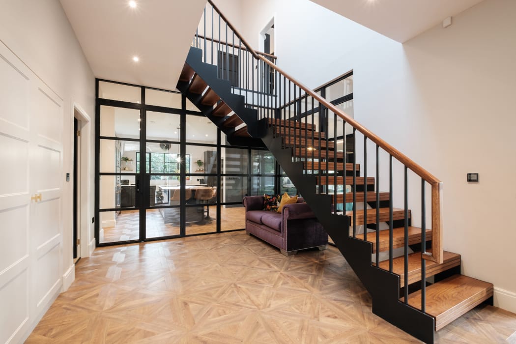 A modern interior with a wooden staircase and metal railing, leading to the upper floor. The space features parquet flooring, a glass wall with doors, and a purple sofa.