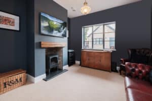 Modern living room with a fireplace, wooden dresser, red armchair, and a wall-mounted TV. Large window with a view of a brick building outside. Beige carpet and dark blue walls.