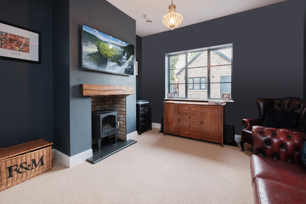 Modern living room with a fireplace, wooden dresser, red armchair, and a wall-mounted TV. Large window with a view of a brick building outside. Beige carpet and dark blue walls.