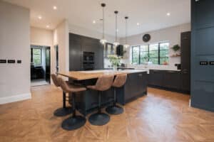 Modern kitchen featuring a large island with wooden countertop, surrounded by dark cabinetry, stools, pendant lights, and a window with a clock above it.