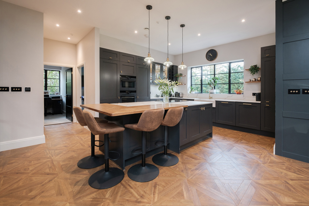 Modern kitchen featuring a large island with wooden countertop, surrounded by dark cabinetry, stools, pendant lights, and a window with a clock above it.