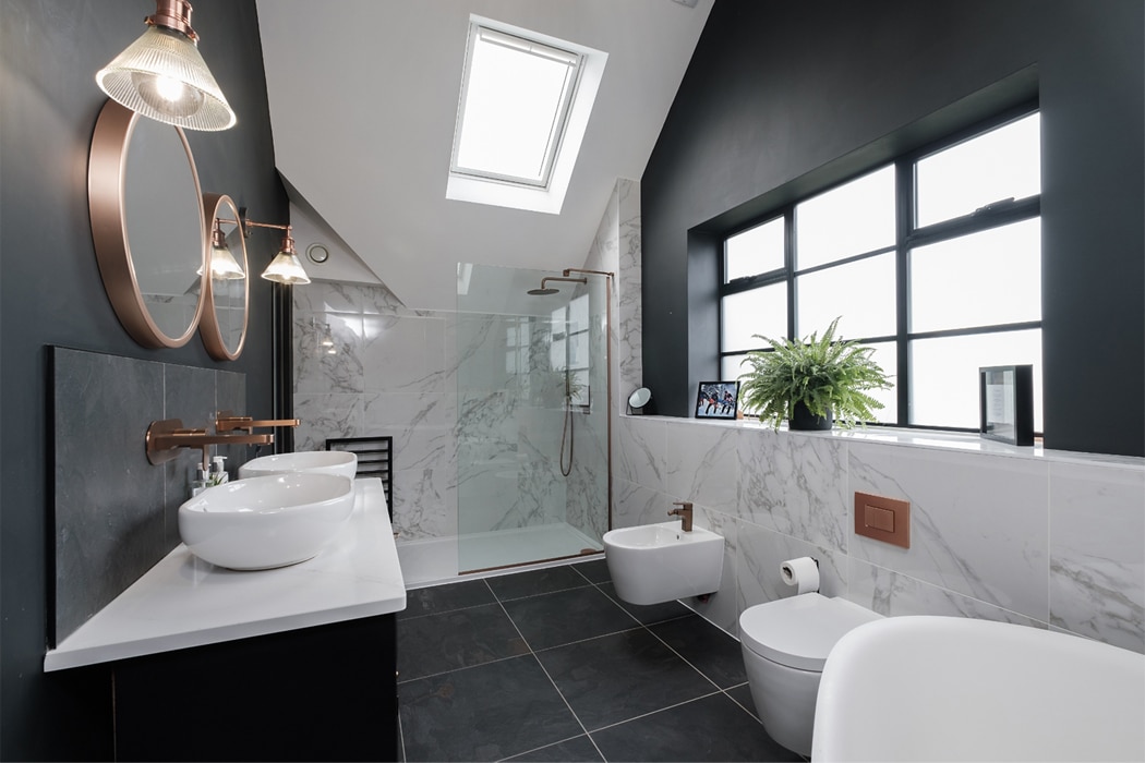 Modern bathroom with skylight, dual sinks, round mirrors, wall-mounted faucets, glass shower, marbled tiles, black accent wall, bidet, and potted plant near a large window.