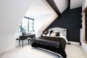 Modern bedroom with angled ceiling, featuring a bed with black and white bedding, a black wall, desk with chair, and a large window.
