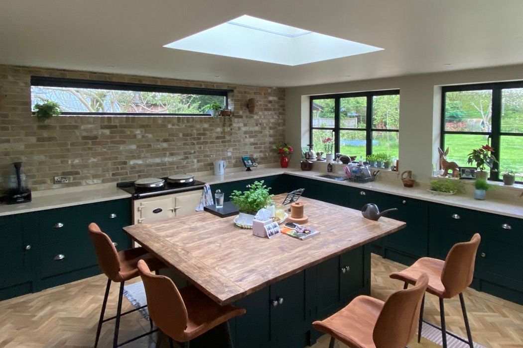A modern kitchen with green cabinets, a wooden island, and brown chairs. Brick walls, large windows, and a skylight provide natural light. Various plants and kitchen items decorate the space.