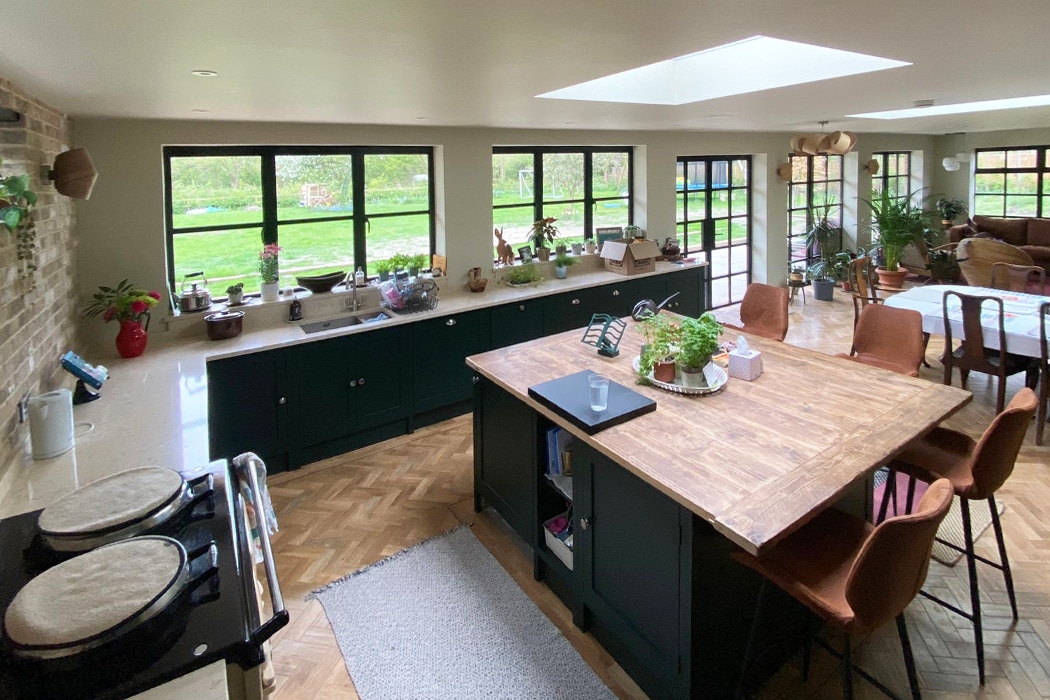 Modern kitchen with a large wooden island, green cabinets, and an adjacent dining area. Windows provide a view of a garden, and plants decorate the space.