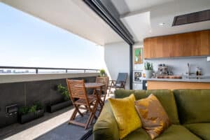 Modern balcony with wooden chairs and a table, overlooking a cityscape. Inside, a cozy kitchen area features wooden cabinets and a green sofa with yellow pillows.